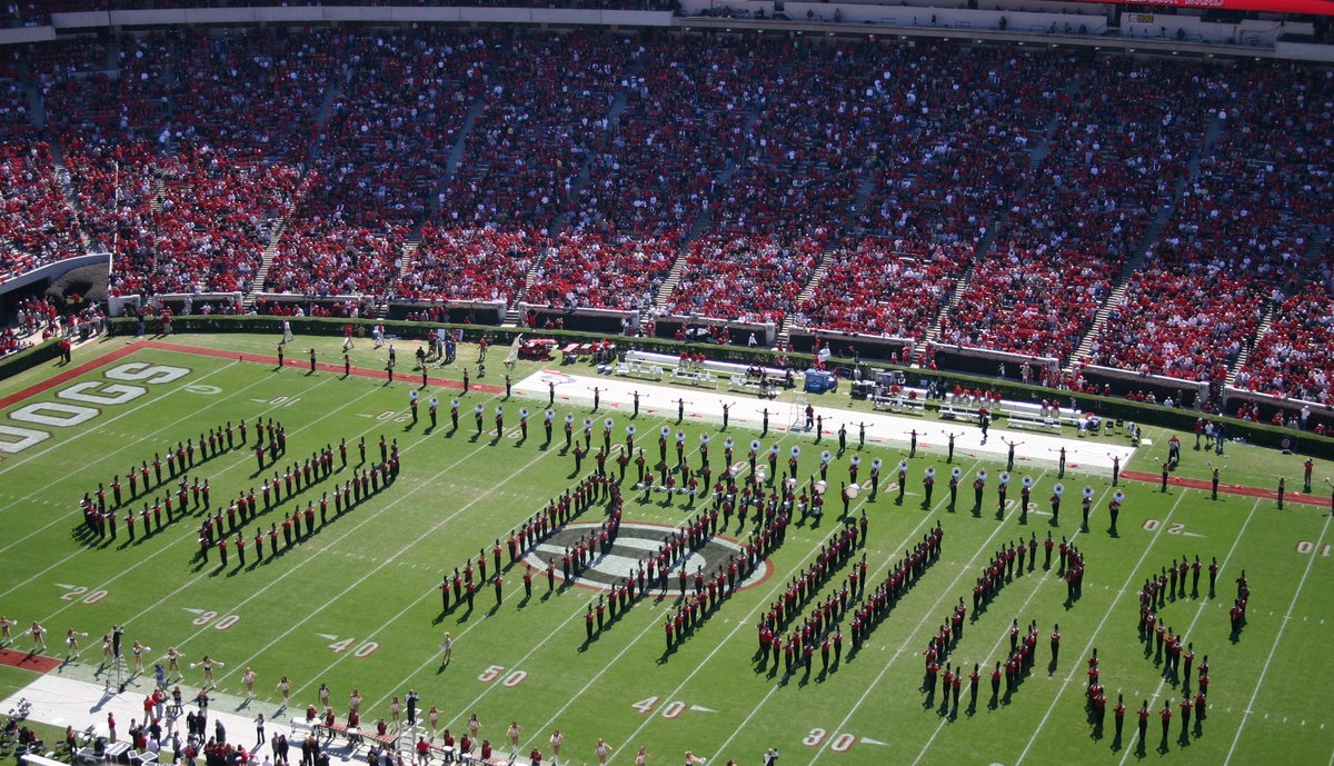 The Redcoat Marching Band in formation on the field at Sanford Stadium, with packed red stands behind them.